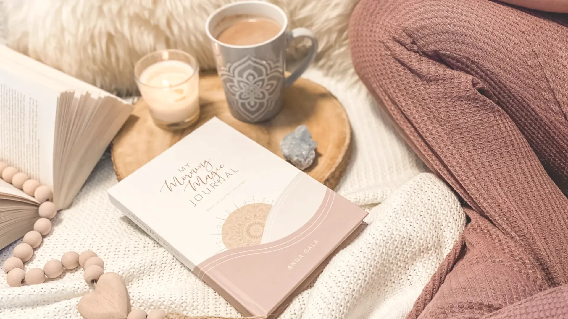 My Morning Magic Journal on a cream knit blanket with a candle, mug of cocoa, wooden beads and a crystal